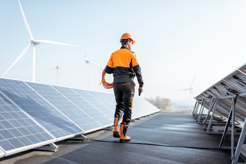 Well-equipped worker in protective orange clothing walking and examining solar panels on a photovoltaic rooftop plant. Concept of maintenance and installation of solar stations